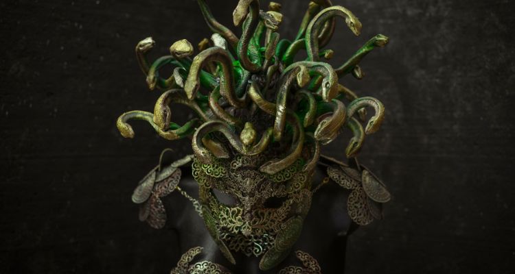 A mask made of green-gold filigreed metal and topped with a gorgon-like headdress of metal snakes rests on the top of a black mannequin wearing matching shoulder pieces.