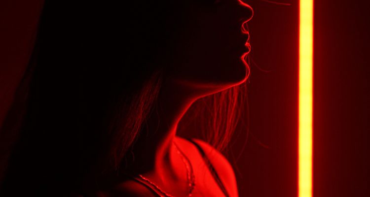 A close up of a woman's upturned lower face, neck, and collarbones backlit by vivid red light while her long hair is in shadow.