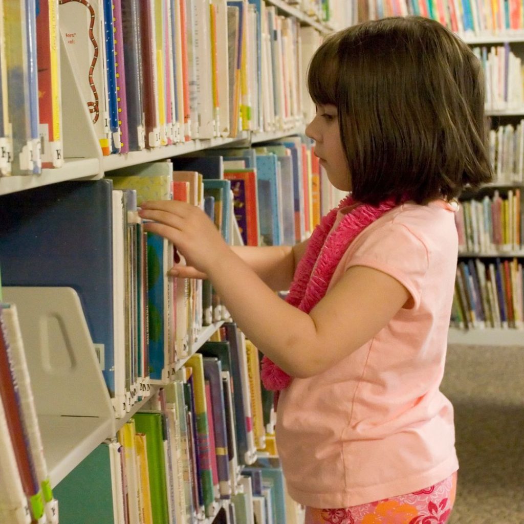 A pale-skinned young child with shoulder-length brown hair browses the spines of picture books on a library shelf.