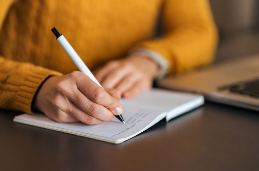 Someone writing in a notebook next to their laptop