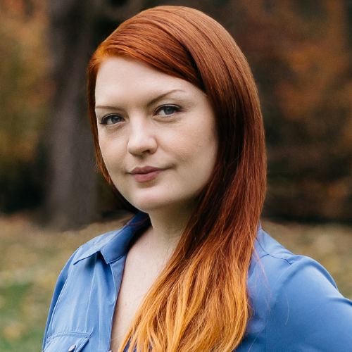 Emily Suvada wearing blue against a wooded background with her red hair to the side.