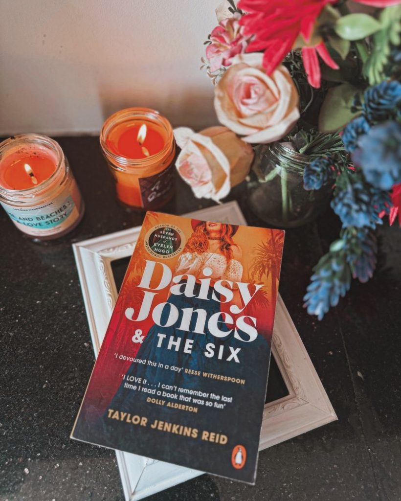 Image by @iambibliomane of a book called "Daisy Jones and the Six" by Taylor Jenkins Reid. The book is next to a vase of flowers and candles.