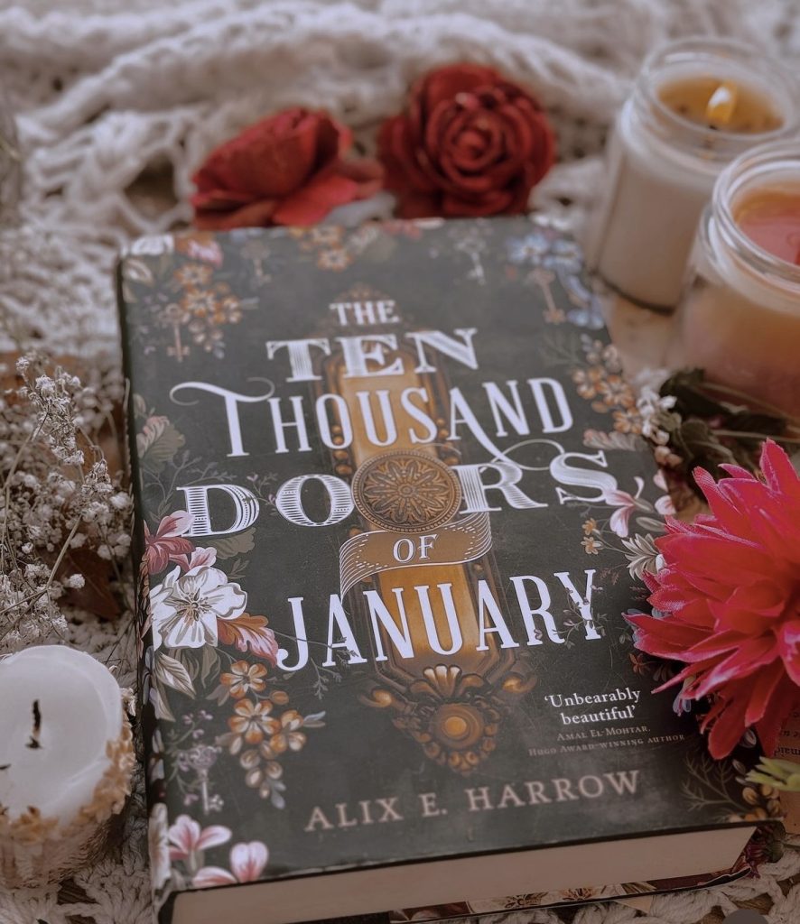 Image provided by @iambibliomane of a book cover of "The Ten Thousand Doors of January" by Alix E. Harrow. The book is surrounded by loose flowers and candles, with one lit.