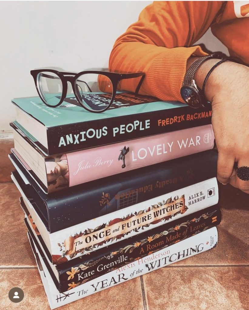 Image by @iambibliomane of a stack of books with Swarna's arm on top of it with a pair of glasses next to her arm.