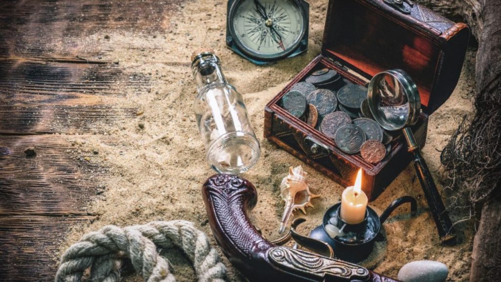 Unveiling The Sensational History Behind Fiction’s Most Famous Pirates By M.A. Cole. It Shows an open treasure box, and other pirate paraphernalia on top of a pile of sand.