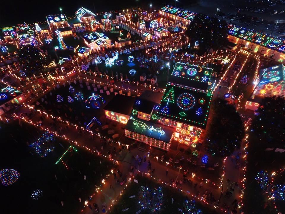 An aerial view of a suburban neighborhood at night, with each home decked out in elaborate, colorful Christmas light displays, including images of stars, snowflakes, bells, and Christmas trees displayed on the roofs.