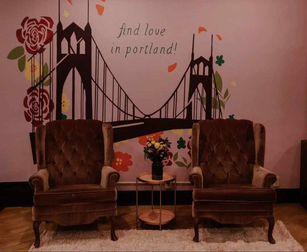 Two brown arm chairs with a table in between with flowers in front of an art mural with a suspended bridge and roses