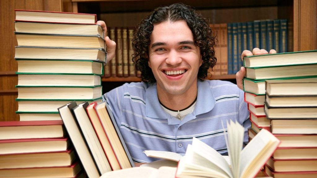 A man is smiling as he peers through tall stacks of books. There is a bookshelf behind him and on the desk in front of him.