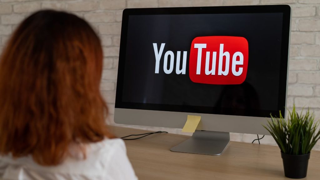 The back of a woman's head can be seen staring at a desktop monitor. The screen has the YouTube logo pulled up.
