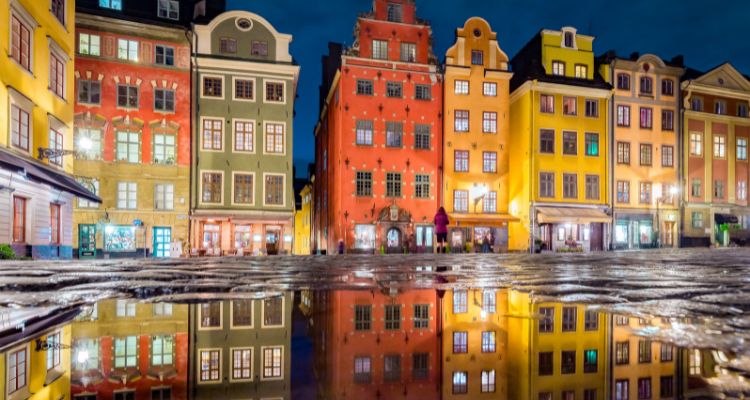 Color building faces with a brick street in the foreground with a puddle reflecting the cityscape