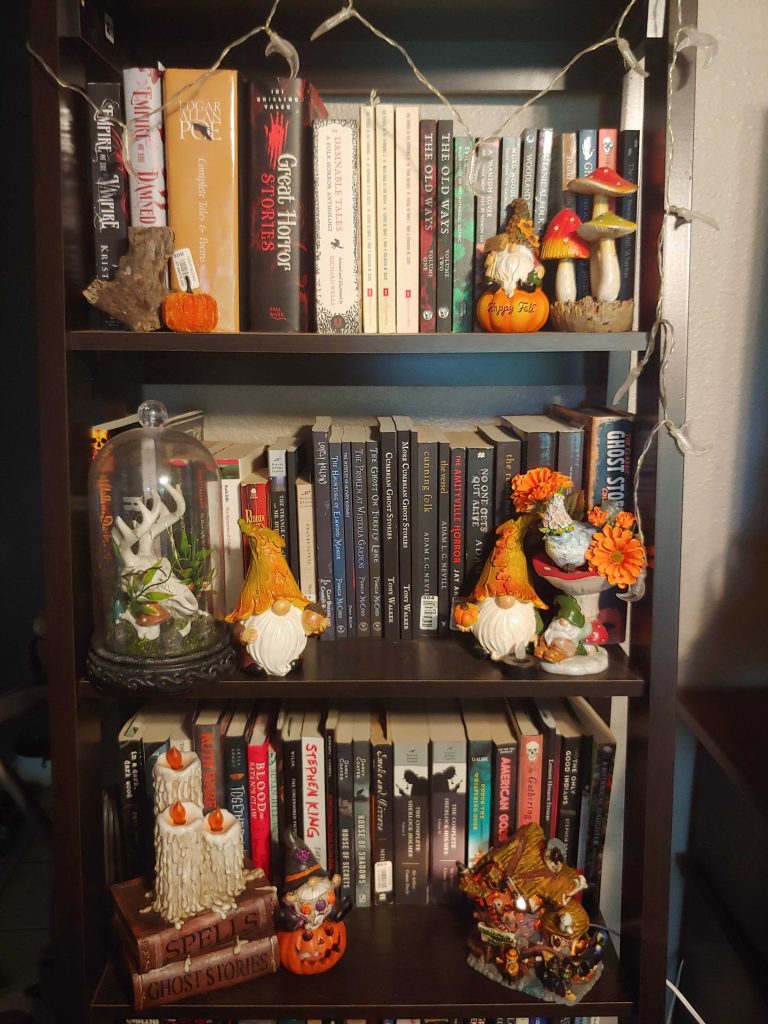 There are two shelfs packed with horror books. It is decorated with moon string lights, orange pumpkins, two white bearded gnomes, and red and green mushrooms. There is a fake, yet realistic white deer skull in a glass case. From Gray O'Dell.