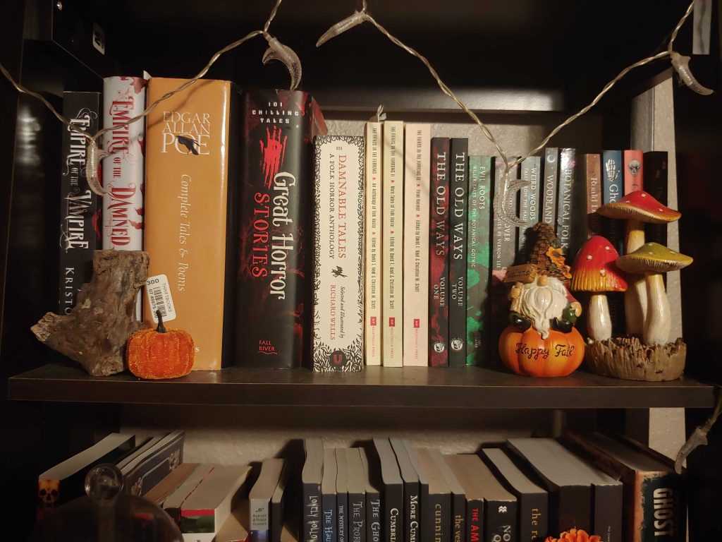A close up of a shelf decorated with moon string lights, orange pumpkins, a white bearded gnome, and red and green mushrooms. There are many organized horror books. From Gray O'Dell.