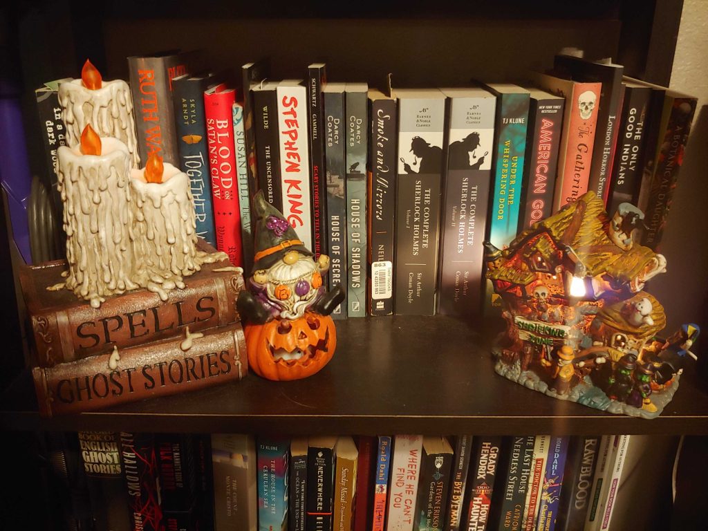 A shelf with many horror books and decor. There are trinkets of two books with three melted candles sitting on top, a purple and orange gnome sitting on a jack-o-lantern, and a little haunted house. From Gray O'Dell.