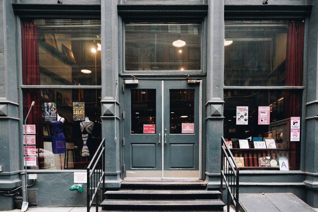 The storefront of Housing Works Bookstore, a set of black steps leading up to pine green double doors surrounded by glass windows with colorful posters hanging up inside.