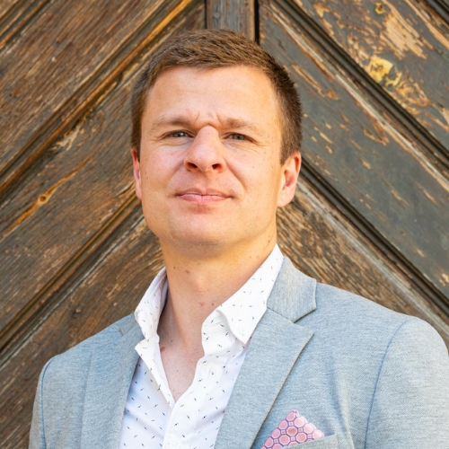 Anthony J. Melchiorri in a gray blazer and printed white shirt in front of a wooden wall.
