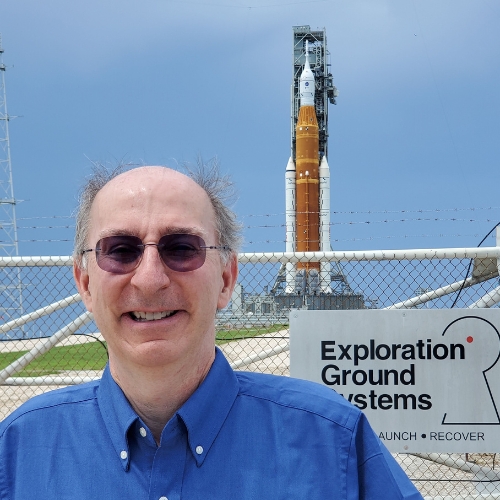 Les Johnson in sunglasses and a blue button-up smiling in front of NASA's Artemis I.