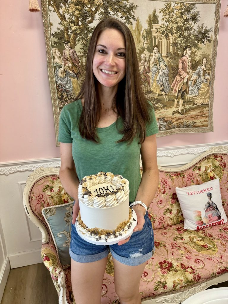 A woman holding a cake that says 10K on the top. She is standing in front of a pink couch covered in flowered with one single pillow. The background is a pink wall with white trimming and a framed painting.