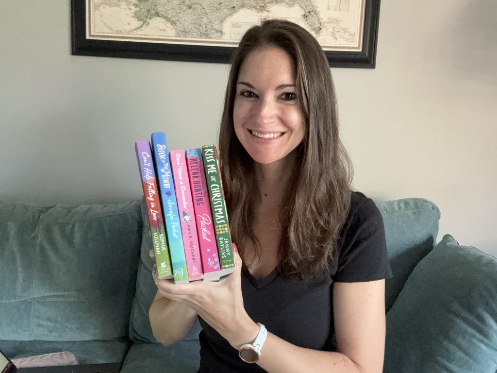 A woman sitting on a green-gray couch, while holding a stack of five books. The couch is placed in front of a cream colored wall with a framed map.