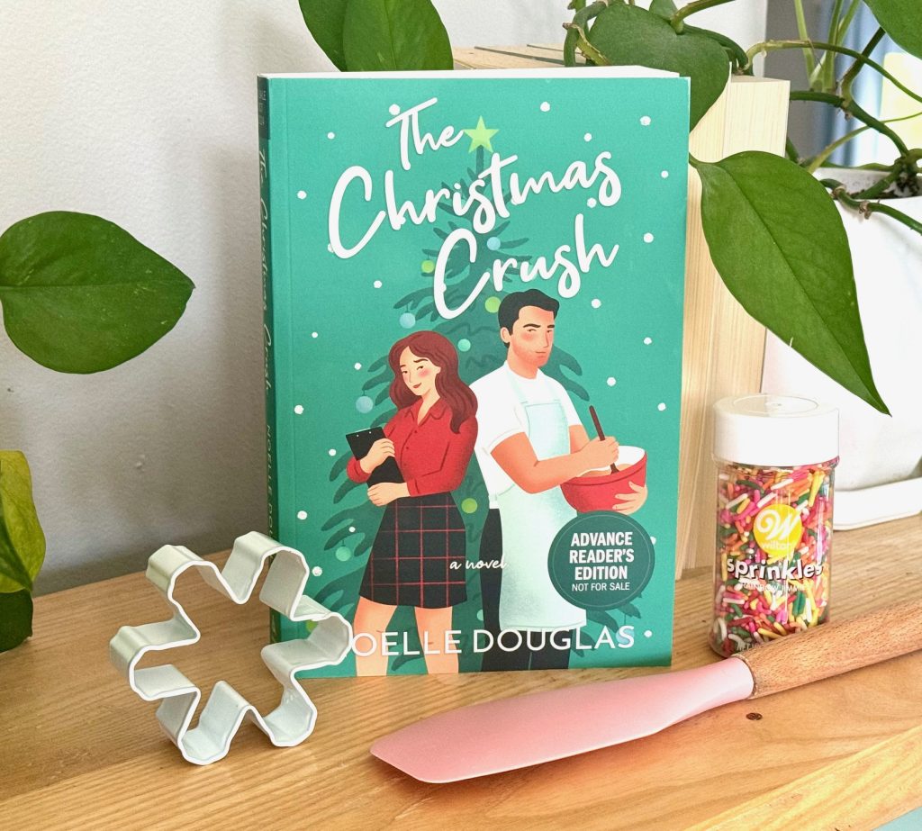 A green book sitting on a wooden shelf. Placed around the book are a plastic spatula, a cookie cutter, and a jar of sprinkles. Two green leafy plants sit on the edge of the frame.