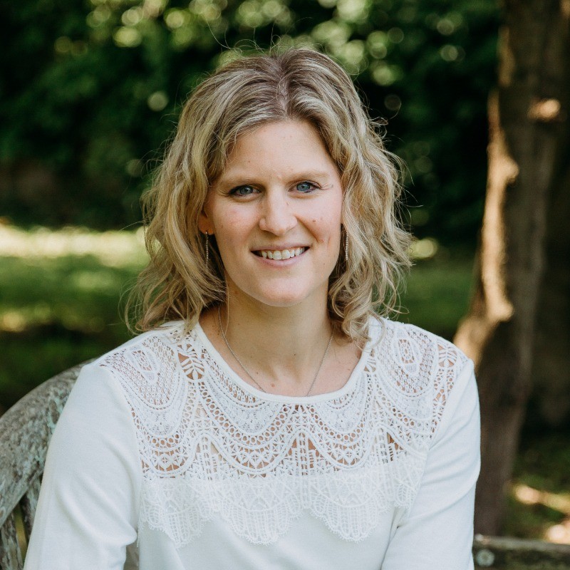 Jessica Correnti sitting outdoors and smiling. She has wavy, shoulder-length blonde hair and wears a longsleeved white shirt with lace trim on the collar.