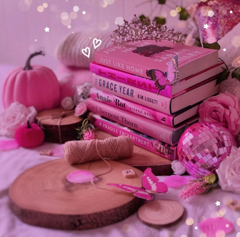 A stack of pink books on a wooden slice, with other wooden slices and rolled-up thread nearby. A tiara sits on top of the books while other pink motifs abound, including butterflies, pumpkins, and a pink crystal ball.