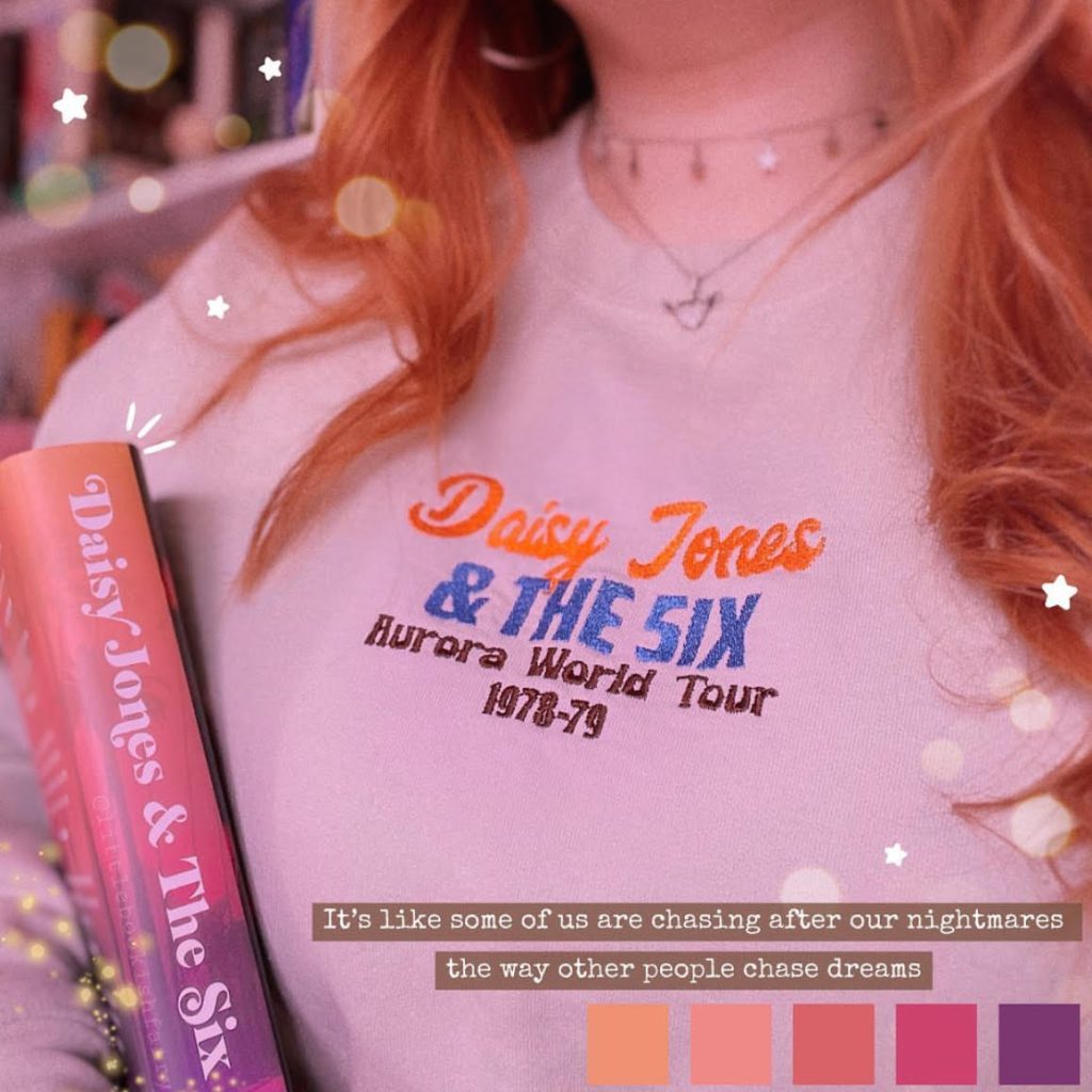 Close-up of an embroidered Daisy Jones & The Six crewneck worn by Lauren. Tucked in her arm is a copy of a book of the same name.