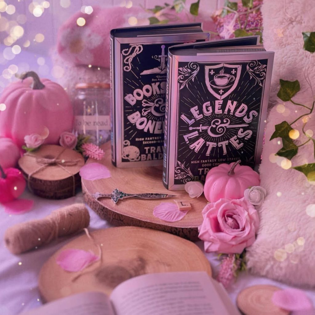 Two hardcover editions of the Legends & Lattes series by Travis Baldree stand vertically on a wooden slice. Surrounding the books are pillows, pink pumpkins and flowers, and more wooden slices.