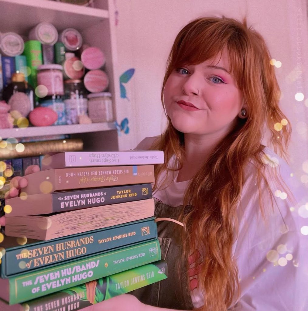 Ginger-haired Lauren holding a stack of multiple editions of The Seven Husbands of Evelyn Hugo by Taylor Jenkins Reid. Behind her is a bookshelf filled with candles; around her are decorative faded lights.