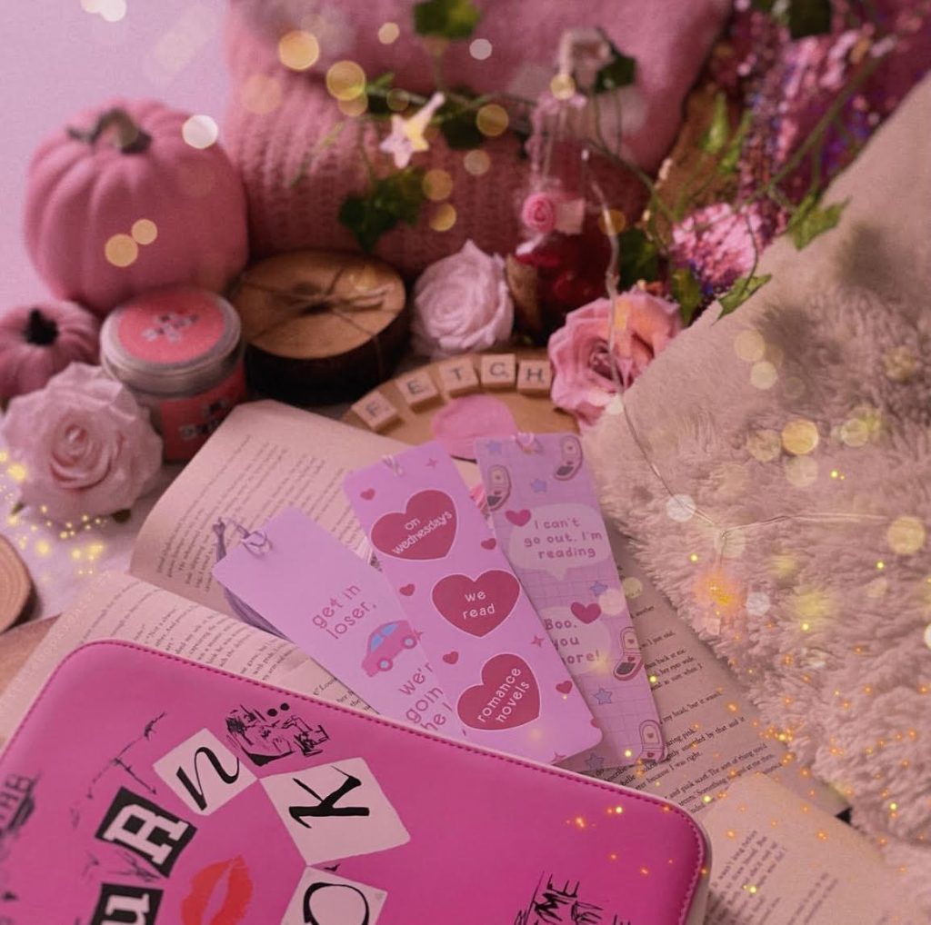 The Mean Girls Burn Book atop a stack of open books and three pink bookmarks. Miscellaneous items surround, including: folded blankets, flowers, pumpkins, and Scrabble letters that spell out FETCH.