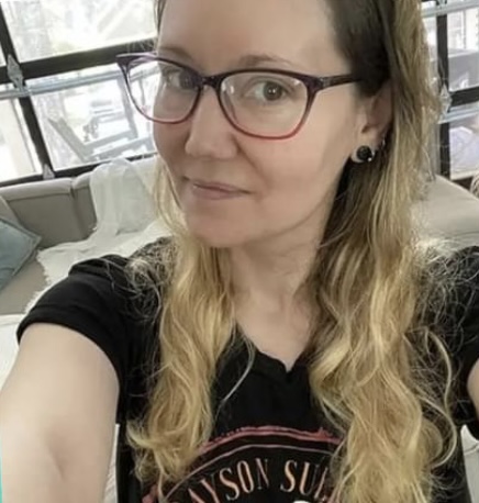 Trisha Wolfe wearing a black t-shirt and red glasses. Her blonde hair is spilling over her shoulders.