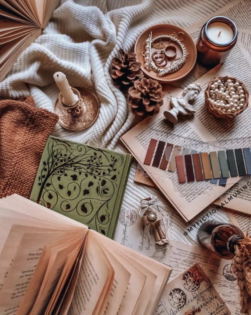A collection of open books, sticky notes, candles, pinecones, and pearl jewelry arranged on top of a white knit blanket.