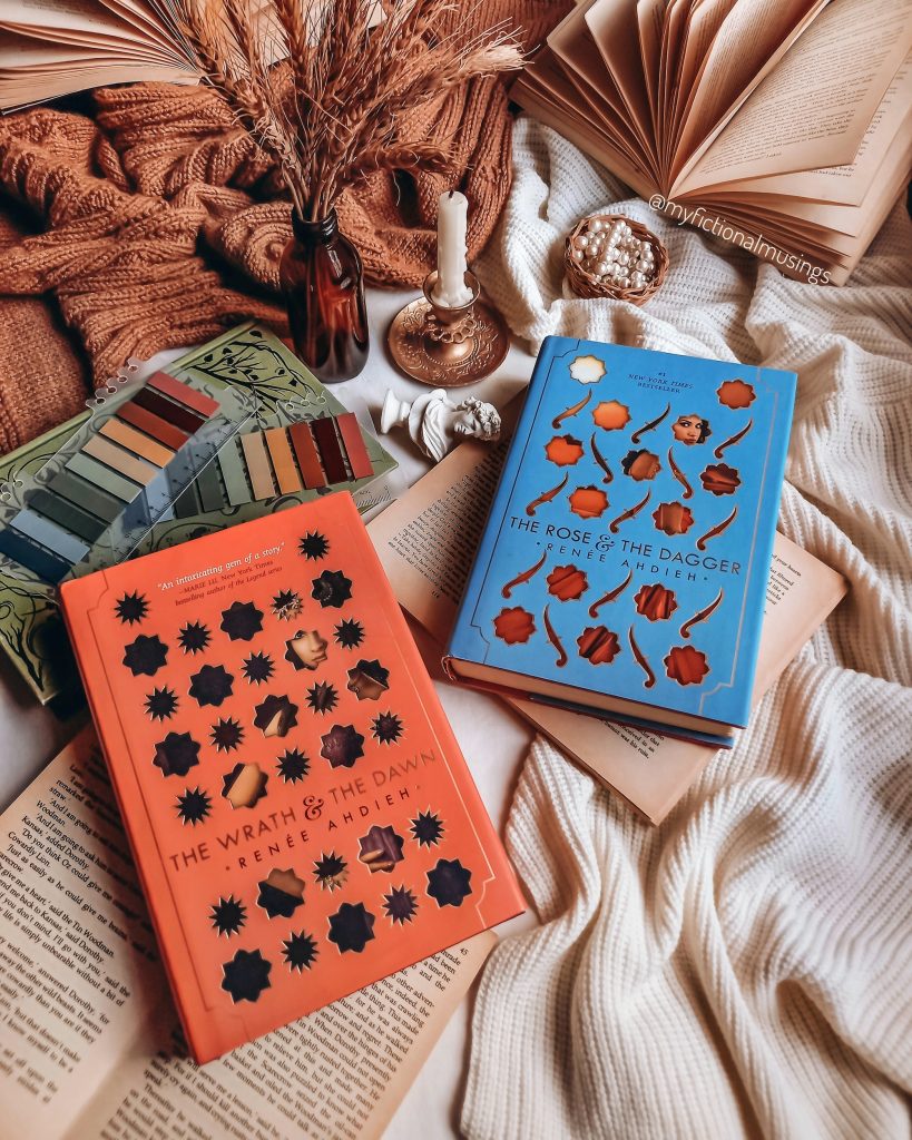 The Wrath and the Dawn and The Rose and the Dagger by Renée Ahdieh lying on top of a white knit blanket next to a melted candle in a gold candle holder and a brown glass bottle holding a few stems of wheat.