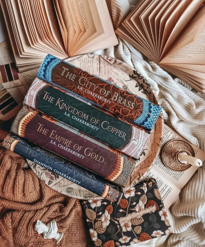 The City of Brass, The Kingdom of Copper, The Empire of Gold, and The River of Silver by S.A. Chakraborty stacked on top of on another, surrounded by other open books with fanned pages and a melted candle in a gold candle holder.
