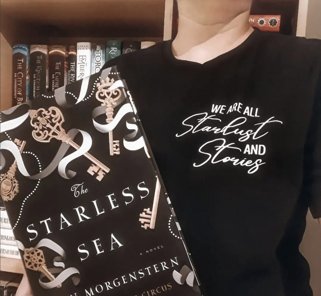 Mitra holding a copy of The Starless Sea by Erin Morgenstern featuring golden keys and white ribbons while wearing a black t-shirt that reads "We are all stardust and stories."