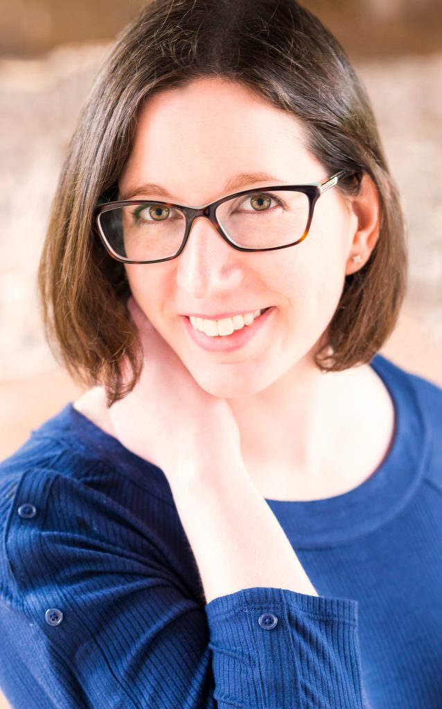 Author Megan Collins smiling while she looks at the camera. Megan has chin-length brown hair and wears a pair of black rectangular glasses and a blue sweater.