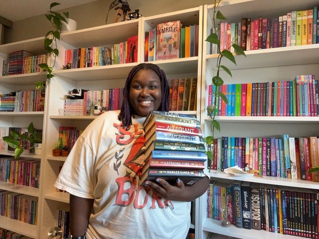 Woman holding a stack of books standing in front of bookcases with green ivies hanging from the shelves