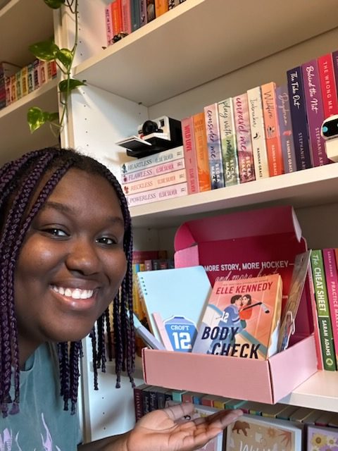 Woman smiling and gesturing toward a pink cardboard box with books on the shelf of a bookshelf