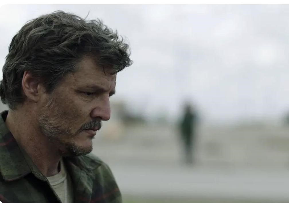 Picture of character Joel from the series The Last of Us wearing a white shirt and green jacket. There is a blurred person in the background.