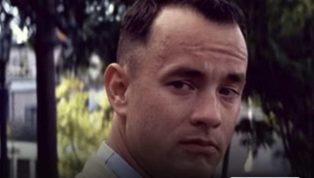 Picture of character Forrest Gump wearing a white shirt with a beige coat in the movie Forrest Gump. There are trees and a green bush in the background.