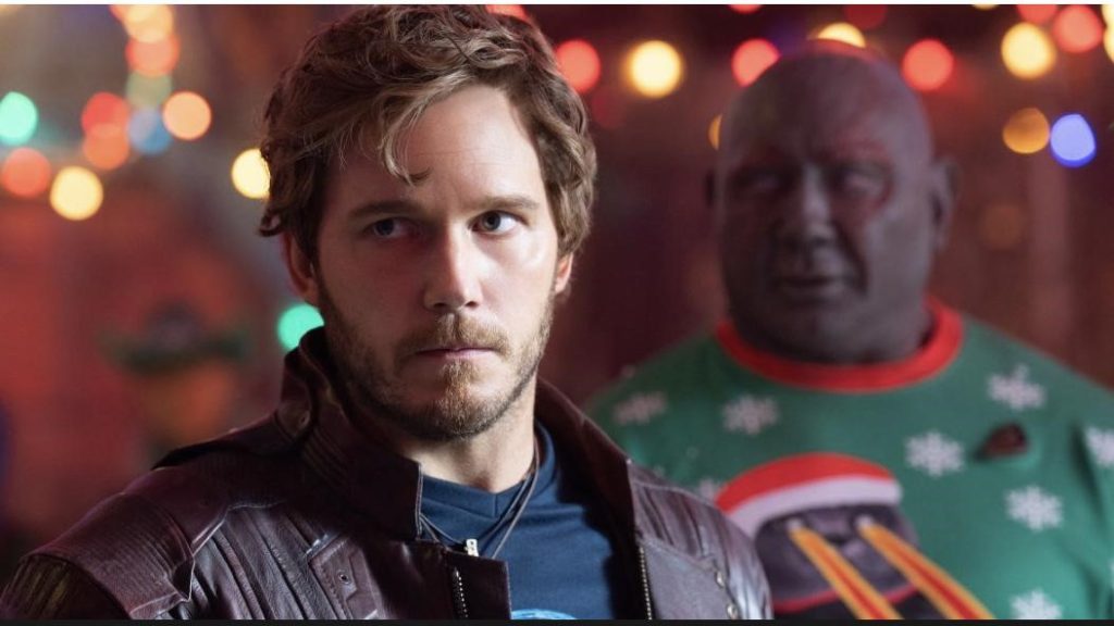 Picture of the character Peter Quill from The Guardians of The Galaxy movie wearing a blue shirt, purple jacket, and a black necklace. Another character, Drax the Destroyer from the same movie, is in the background. And there are yellow and red lights around them.
