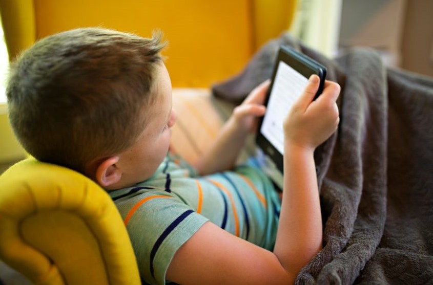 A boy lying on the couch and reading off a Kindle.
