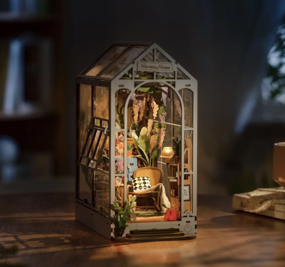 Small wooden book nook that looks like a greenhouse. Inside are a variety of plants, a lamp, a small corner bookshelf, and a wicker chair with a pillow and blanket. The greenhouse nook has a very cozy vibe.