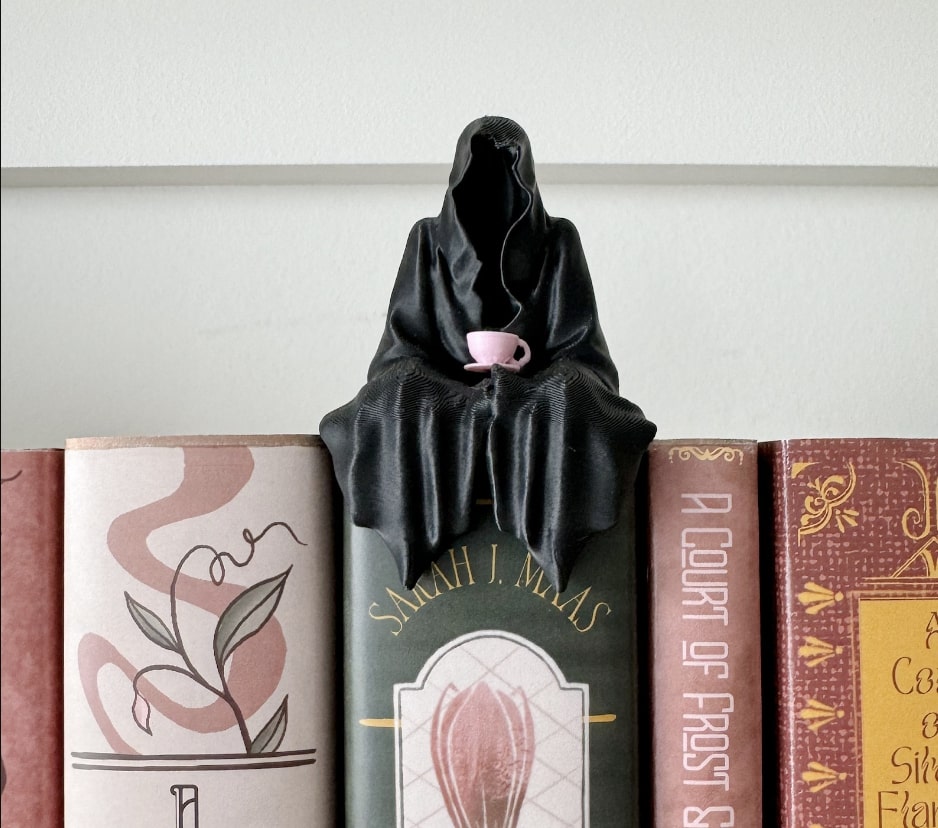 A grim reaper shrouded in a black cloak with no face holds a pink teacup and saucer on its lap. It is about the width of a thick chapter book and sits on the edge of a row of books.