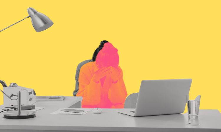 Woman holds her head in her hands behind a desk.