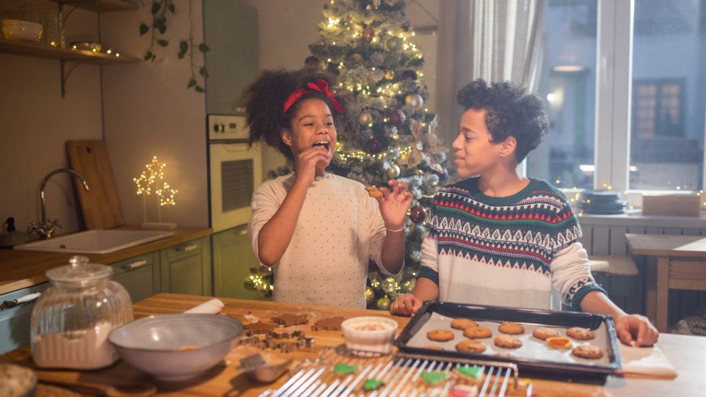 Two kids baking cookies the girl eats one and the boy watches. A big Christmas tree is behind them by the oven.