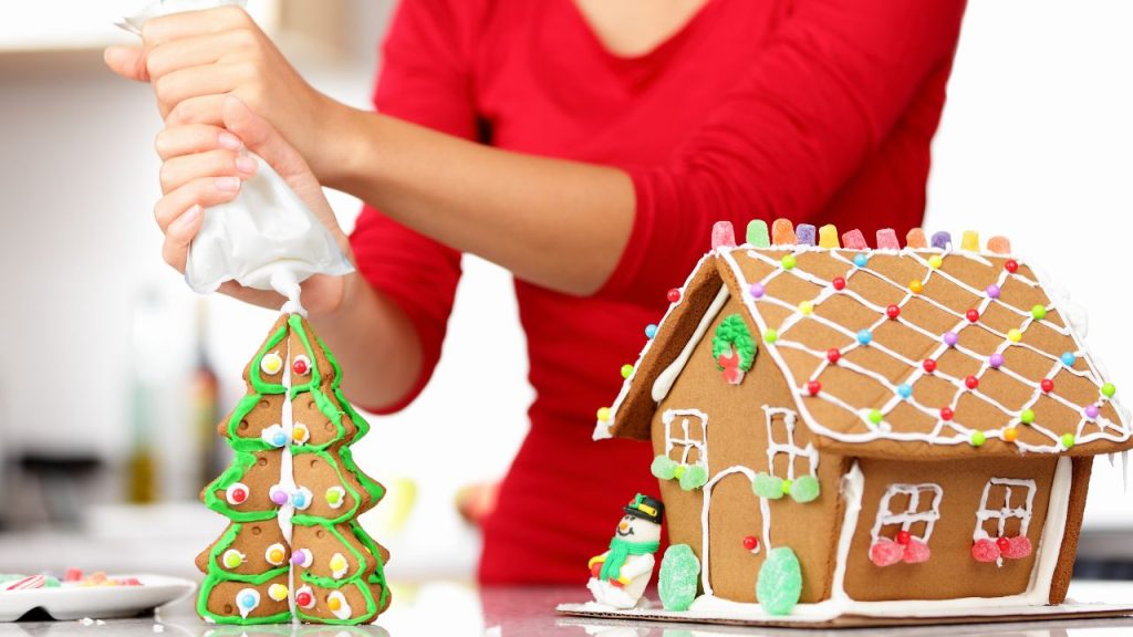 Gingerbread house with a snowman leaning on the decorated house as a woman pipes icing on the top of a Christmas tree gingerbread.