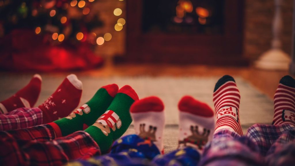 Four pairs of feet with Christmas-themed socks as the family sits on the floor in front of a fireplace and Christmas tree.
