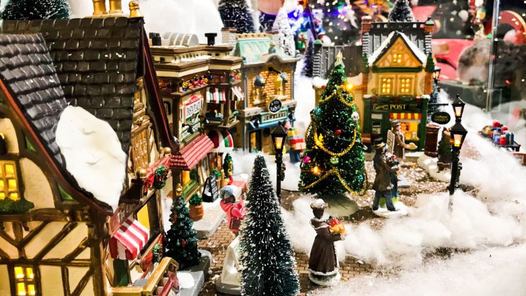 A Christmas small figurine town of different stores and a Christmas tree in the middle of the town. Cotton fluff is spread throughout like snow.