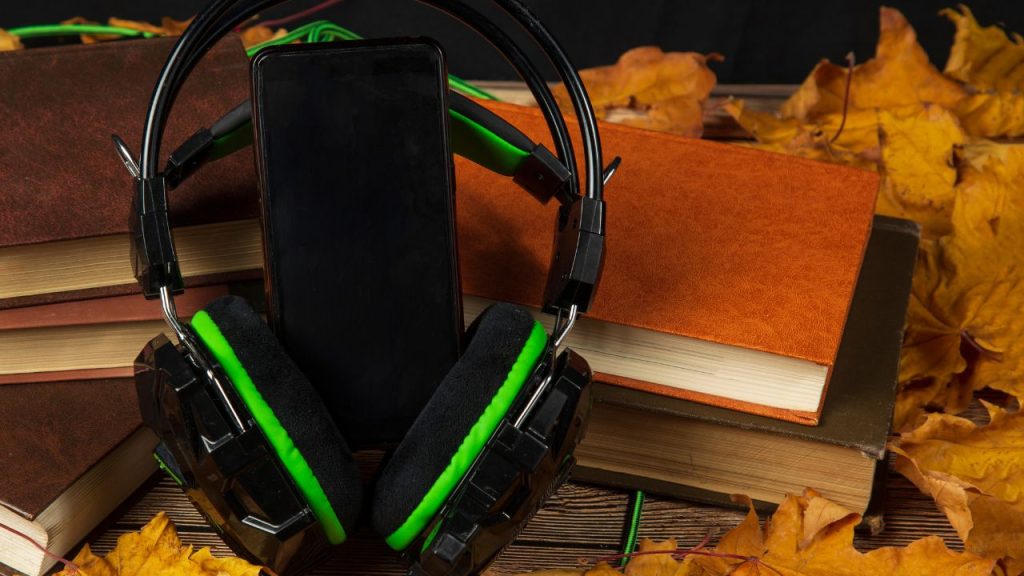 A close-up of a black phone and a set of black and neon green headphones leaning on a pile of brown notebooks surrounded by yellow leaves.