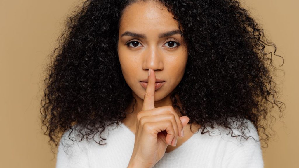 A close-up of a young Black woman with curly black hair holding up a finger to her mouth in a shushing motion.